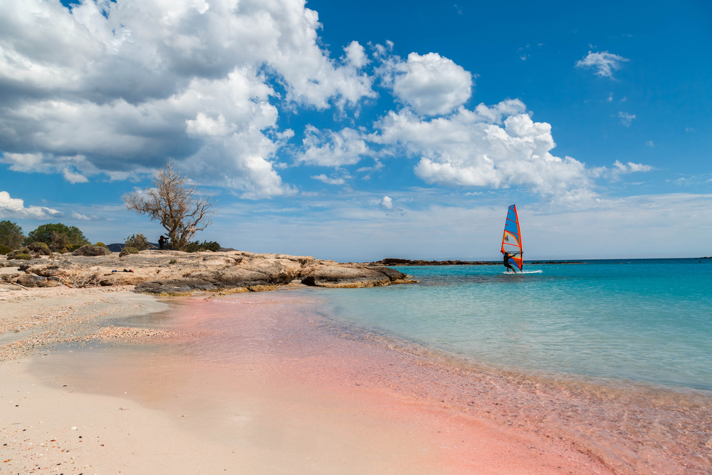 Windsurfař klouže po azurovém moři u růžové písčité pláže v Řecku, obklopen krásnou přírodní krajinou a jasným modrým nebem.