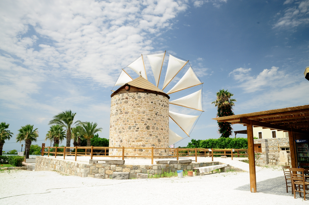 Tradiční kamenný mlýn s bílými lopatkami v jasně osvětlené krajině ostrova Kos, obklopený palmami a jasně modrou oblohou, typická řecká architektura na Hippokratově ostrově