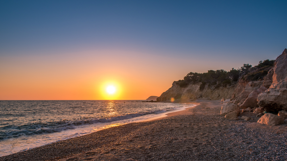 Západ slunce na řecké oblázkové pláži s vlnami omývajícími pobřeží a světlo slunce odrážející se od hladiny moře, klidný večerní pohled ideální pro návštěvníky