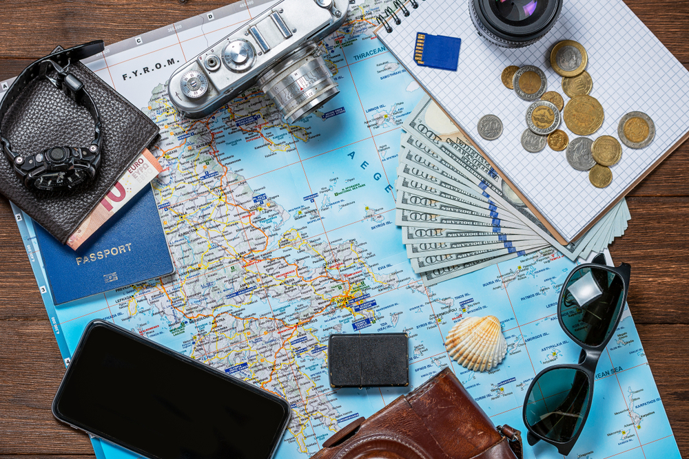 Mapa Řecka obklopená cestovními potřebami, jako je fotoaparát, peníze, mince, zápisník, hodinky, peněženka s eury, pas, smartphone, sluneční brýle a mušle.
