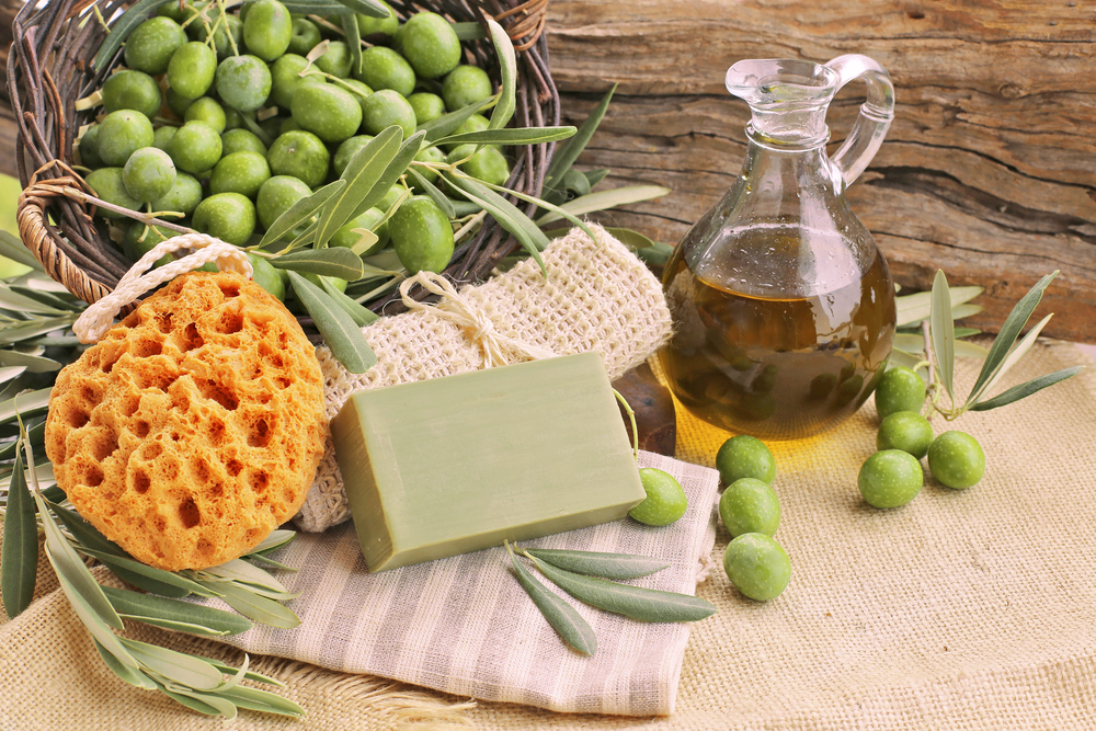 Památky z Řecka – co si přivézt ze země tisíce ostrovů? 3 Košík s čerstvými zelenými olivami, olivový olej v karafě, přírodní houba a olivové mýdlo na rustikálním pozadí.