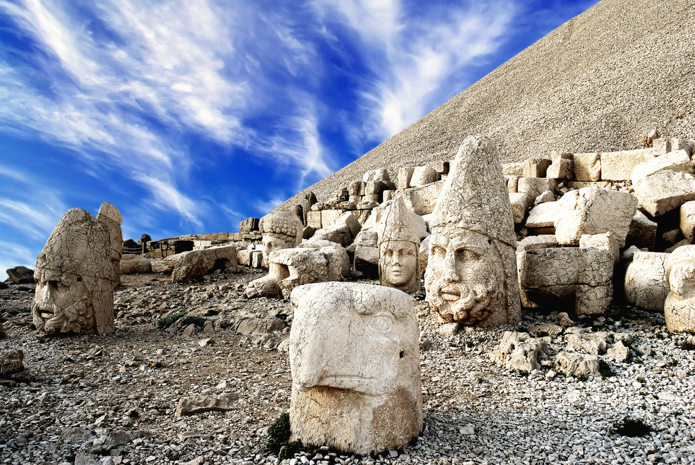 Sochy kamenných hlav na hoře Nemrut, obklopené kamenitým terénem, pod jasnou modrou oblohou s bílými mraky.