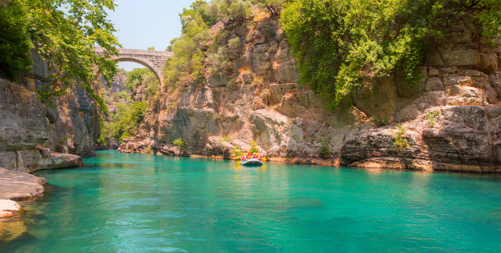 Krásný kaňon s tyrkysově modrou řekou, obklopený strmými skalami a zelenými stromy, s historickým kamenným mostem v pozadí. Skupina lidí na raftu si užívá dobrodružství na řece. Ideální místo pro rodinnou dovolenou s dětmi v Turecku.