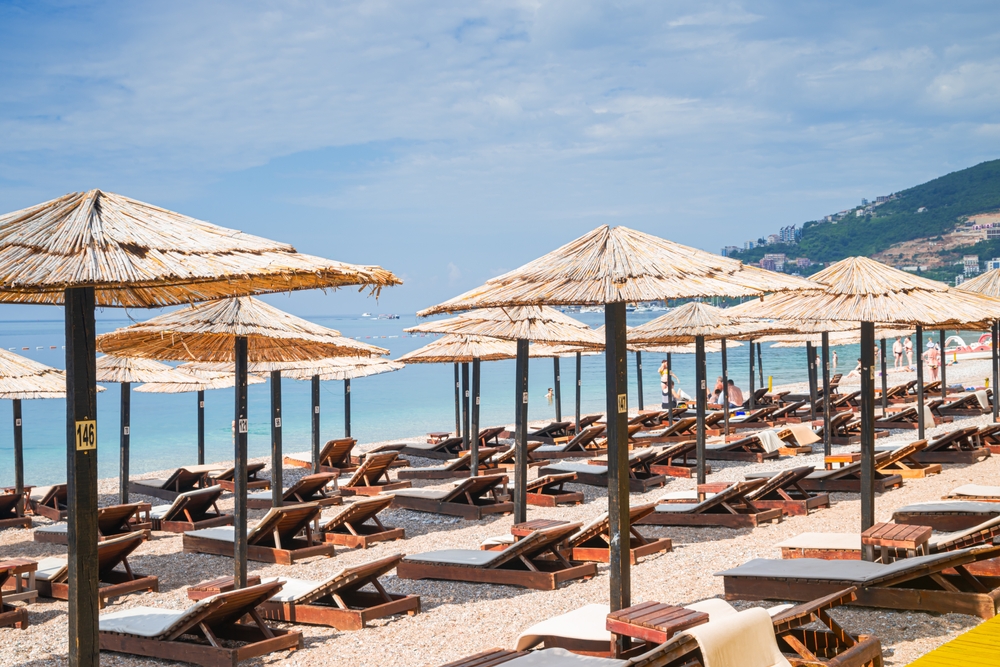 Pláž v Černé hoře se slaměnými slunečníky, dřevěnými lehátky a světle modrým mořem