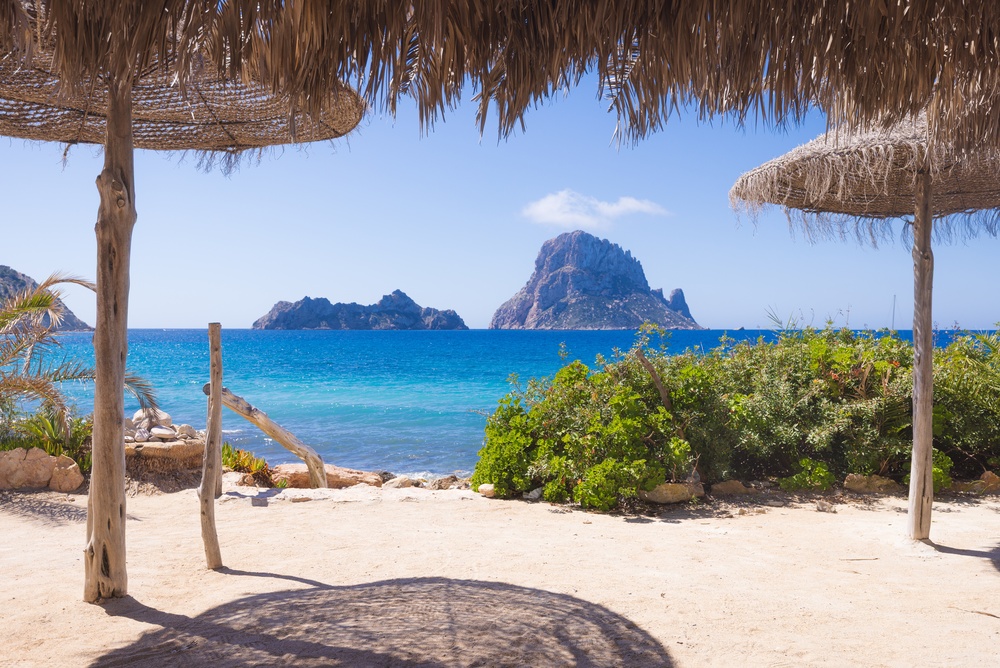 Pláž Cala d’Hort na Ibize se slaměnými slunečníky, zelení a modrým mořem