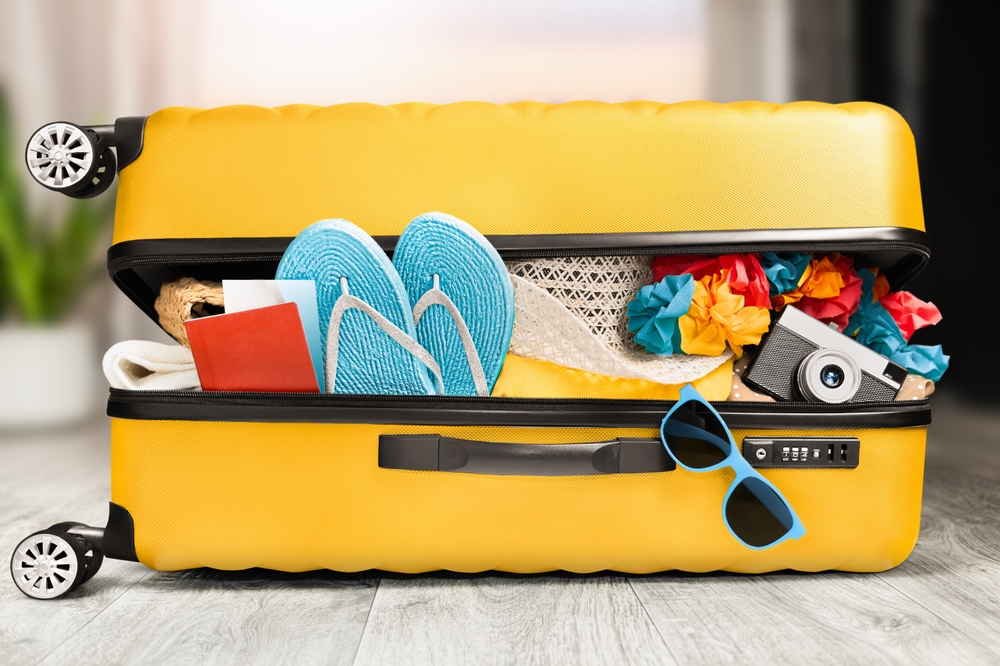 Žlutý kufr sbalený na dovolenou se slunečními brýlemi, žabkami, kloboukem a fotoaparátem