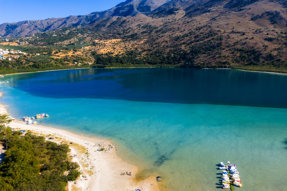 Letecký pohled na jezero Kournas na Krétě v Řecku s tyrkysovými vodami, písečnou pláží s loděmi, drsnými kopci a vzdálenými budovami